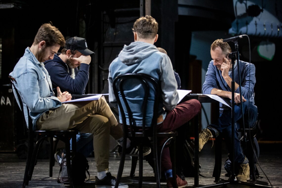 Próba spektaklu na scenie teatru - mężczyźni siedzący przy stole wpatrzeni w scenariusze