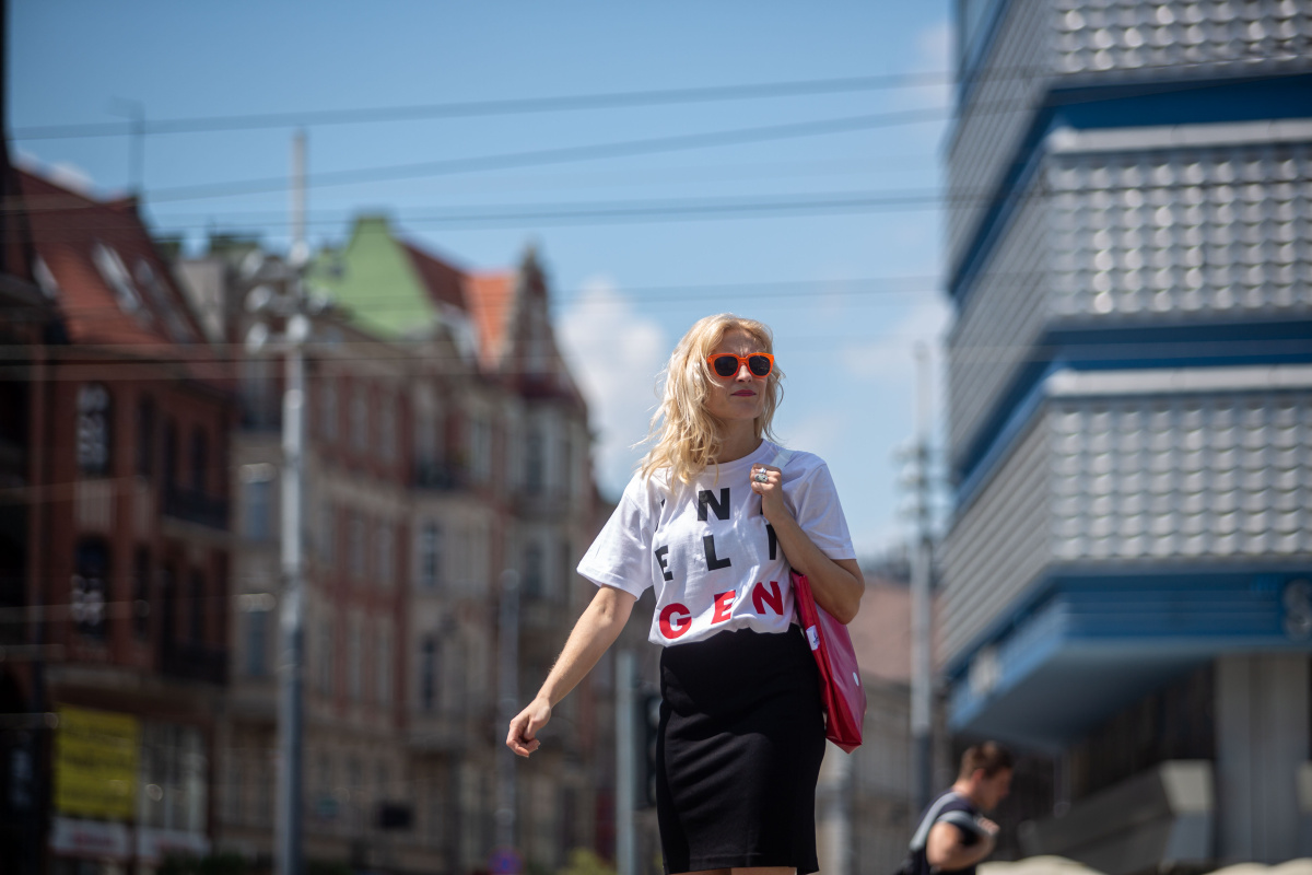 Kobieta na tle miasta w koszulce z napisem Inteligenci, na ramieniu ma różową torbę.