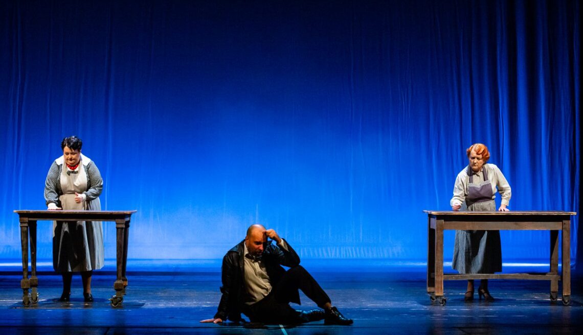 Aktor siedzi na scenie, za nim przy dwóch stołach stoją aktorki.