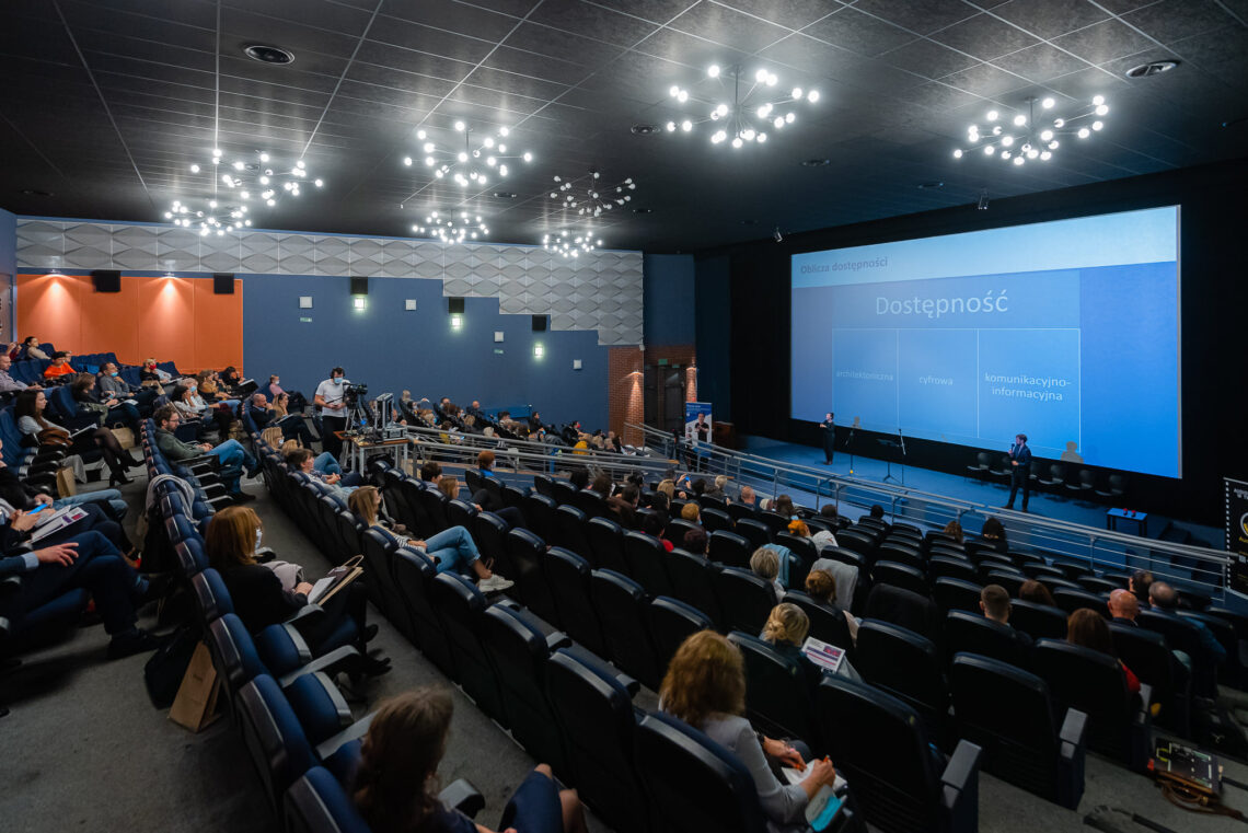 Oświetlona sala kinowa z niebieskimi fotelami, na ekranie wyświetlona prezentacja "Oblicza dostępności"