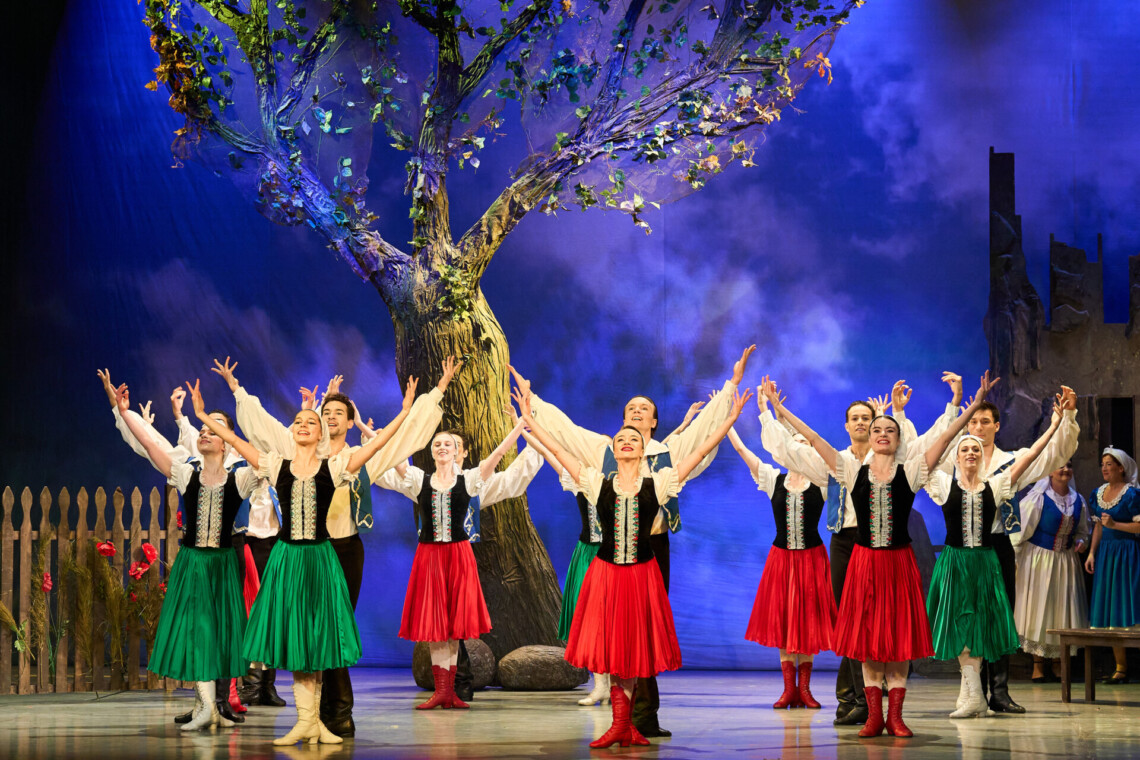 Scena teatralna z dużym drzewem pośrodku, przed którym na pierwszym planie stoją kobiety w czerwonych i zielonych spódnicach z uniesionymi ramionami, między nimi ustawieni są mężczyźni w jasnych koszulach