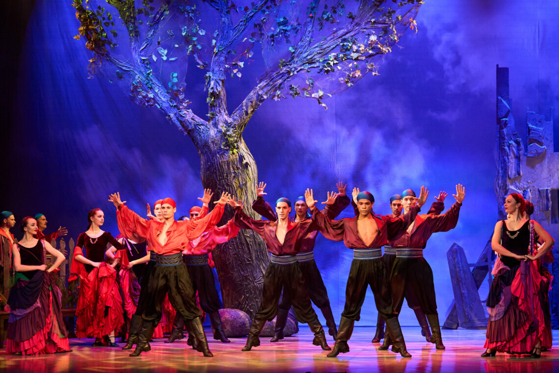 Mężczyźni w czerwonych koszulach i ciemnych spodniach stoją przodem w rzędach z uniesionymi w górę dłońmi, za nimi stoi duże drzewo, po bokach mężczyzn stoją kobiety w sukniach