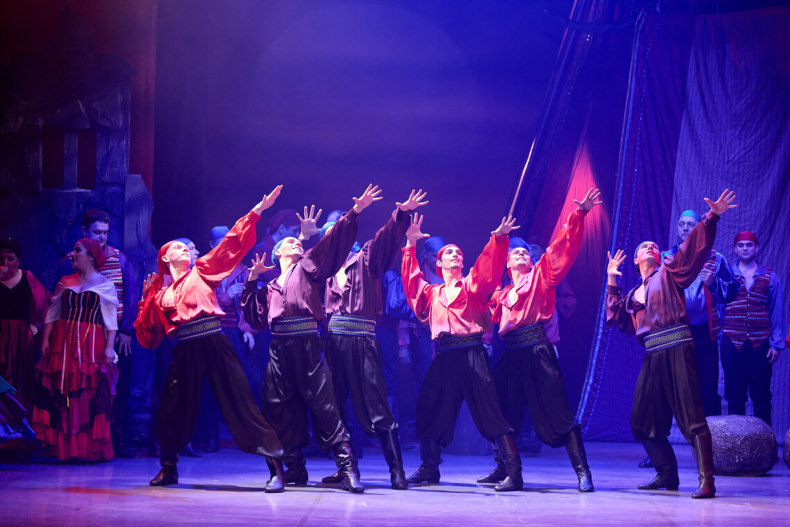 Mężczyźni w czerwonych koszulach i ciemnych spodniach stoją lekko bokiem i pod kątem, z ciałami nachylonymi w jednym kierunku oraz uniesionymi ku górze rękami