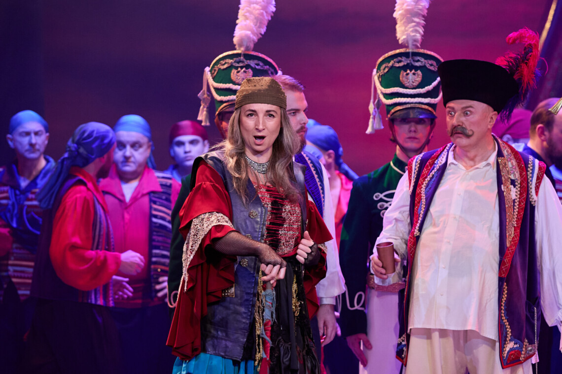 Na pierwszym planie kobieta w czerwonej bluzce i fioletowej kamizelce, zwrócona lekko bokiem z otwartymi ustami, obok niej mężczyzna w jasnym stroju i czapce trzyma kubek, a za nimi gęsto ustawieni są aktorzy w barwnych, wojskowych i ludowych kostiumach na tle purpurowo oświetlonej sceny