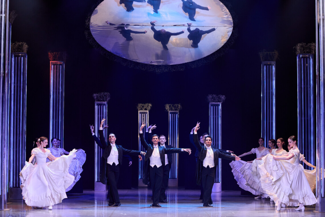 Elegancka scena baletowa z kolumnami, w centrum mężczyźni w czarnych frakach z uniesioną w górę prawą ręką, po bokach kobiety w białych, falbaniastych sukniach, nad nimi okrągłe lustro odbijające sylwetki mężczyzn z góry