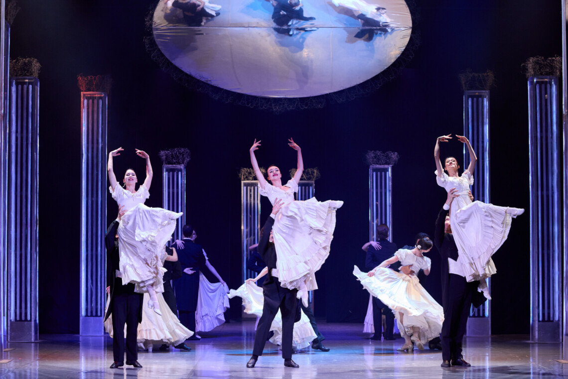 Elegancka scena baletowa z wysokimi kolumnami, mężczyźni w czarnych frakach unoszą partnerki w białych, falbaniastych sukniach, nad nimi znajduje się okrągłe lustro odbijające sylwetki tancerzy z góry