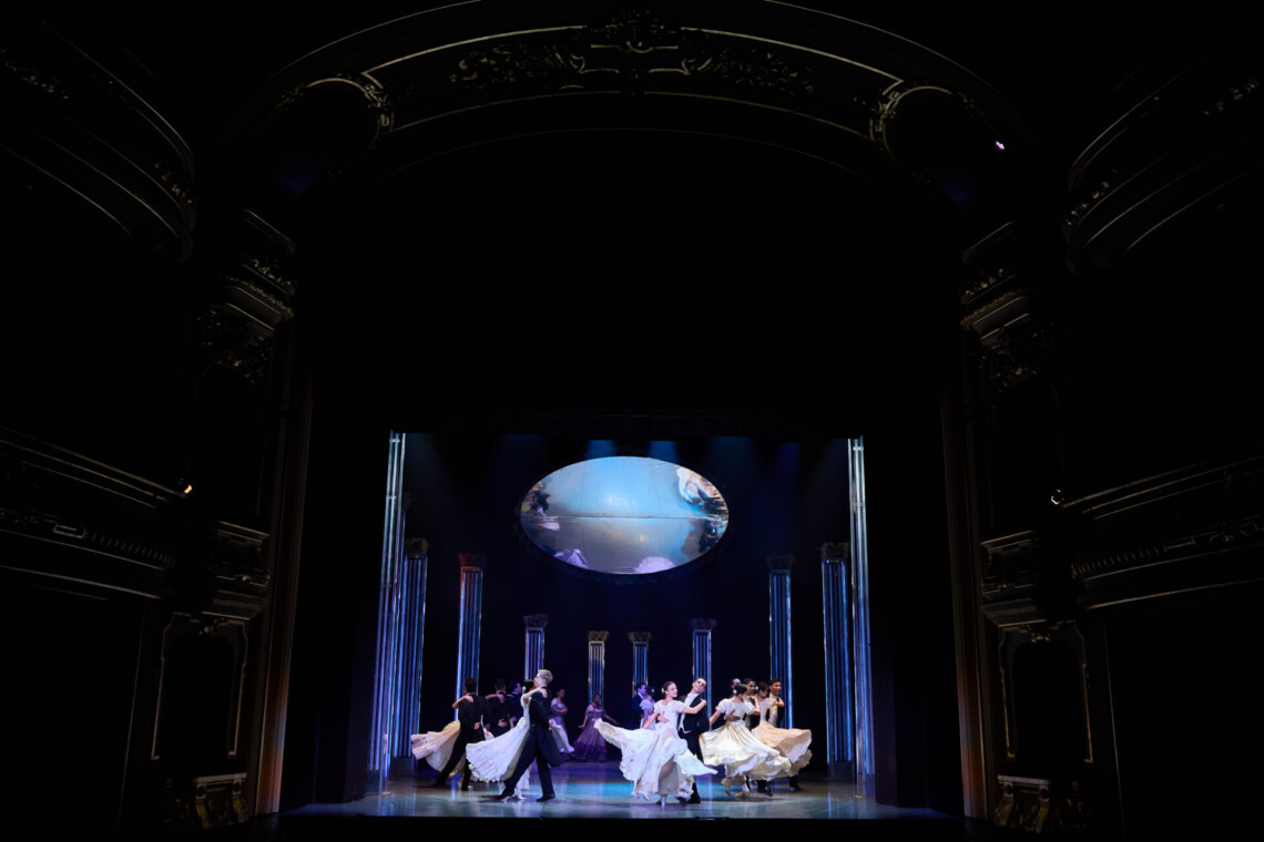 Elegancka scena baletowa z oddali, pary tancerzy w czarnych frakach i jasnych, wirujących sukniach tańczą pomiędzy wysokimi kolumnami, a nad nimi zawieszone jest okrągłe lustro odbijające fragmenty ruchu z góry