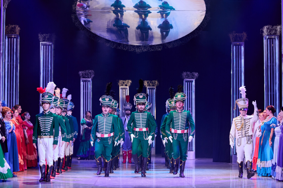 Mężczyźni w zielonych, wojskowych mundurach maszerują środkiem sceny pomiędzy rzędami kobiet w kolorowych sukniach, obok nich wyróżnia się żołnierz ubrany na biało, nad sceną znajduje się duże, okrągłe lustro odbijające sylwetki żołnierzy z góry.