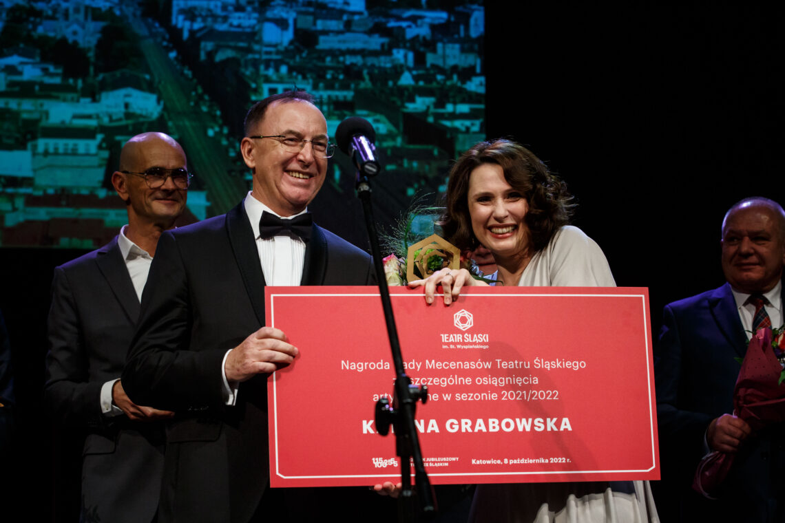 Uśmiechnięta Karina Grabowska z nagrodą Rady Mecenasów Teatru Śląskiego za szczególne osiągnięcia artystyczne w sezonie 2021/2022, w ręce trzyma również statuetkę i kwiaty, obok niej stoi przedstawiciel Rady Mecenasów