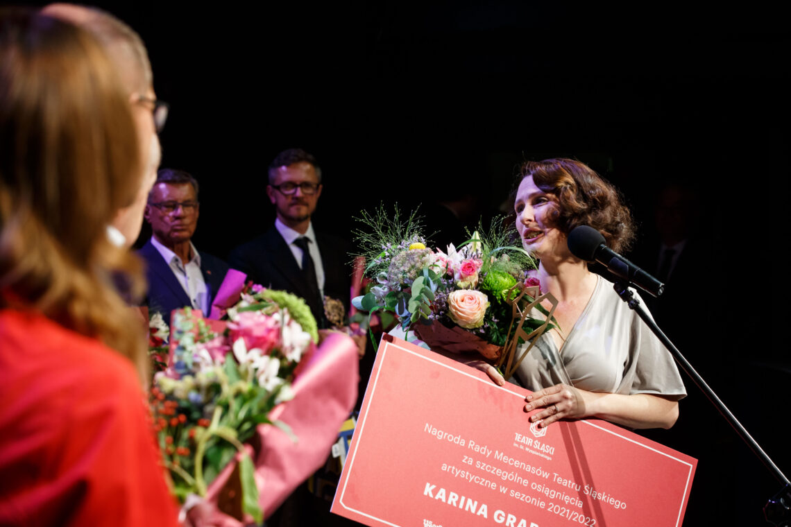 Karina Grabowska przemawiająca do mikrofonu na statywie, odwrócona jest bokiem do osób stojących z jej prawej strony, w ręce trzyma nagrodę Rady Mecenasów - duży prostokątny karton z białymi napisami na czerwonym tle oraz statuetkę i kwiaty