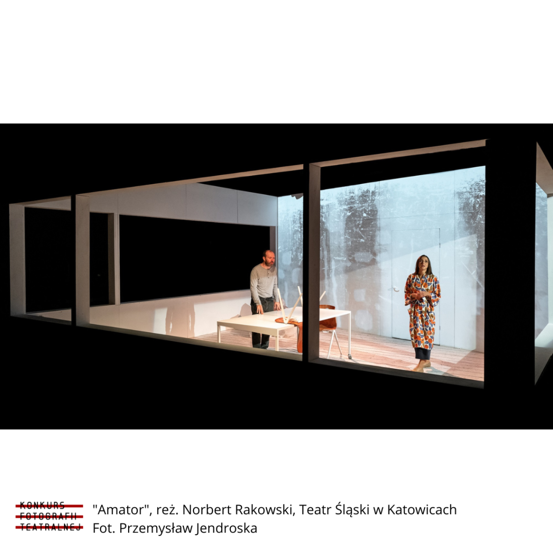 Kadr ze spektaklu Amator 2020: widok z zewnątrz do wnętrza pokoju, w centrum prostokątny stół, przy którym stoi mężczyzna, z prawej strony stoi kobieta ubrana na kolorowo