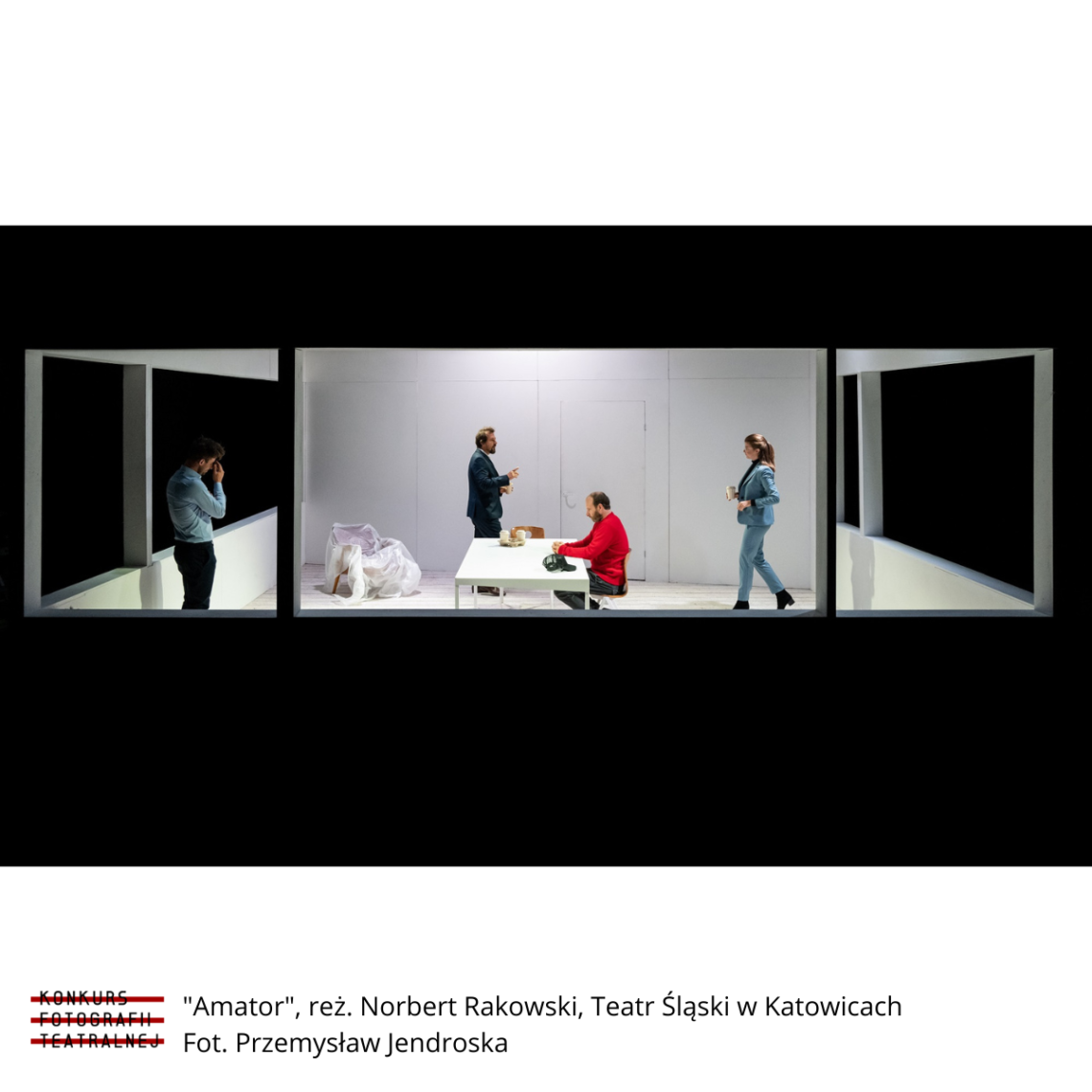 Kadr ze spektaklu Amator 2020: widok z zewnątrz do wnętrza pokoju, w centrum biały prostokątny stół, przy którym siedzi mężczyzna w pomarańczowym swetrze, kobieta i mężczyzna przechadzają się po pomieszczeniu, z boku samotnie stoi mężczyzna