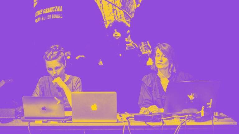 Dwie kobiety siedzące przy stole, przed każdą z nich laptop oraz jeden dodatkowy - zdjęcie w barwach fioletu i żółci