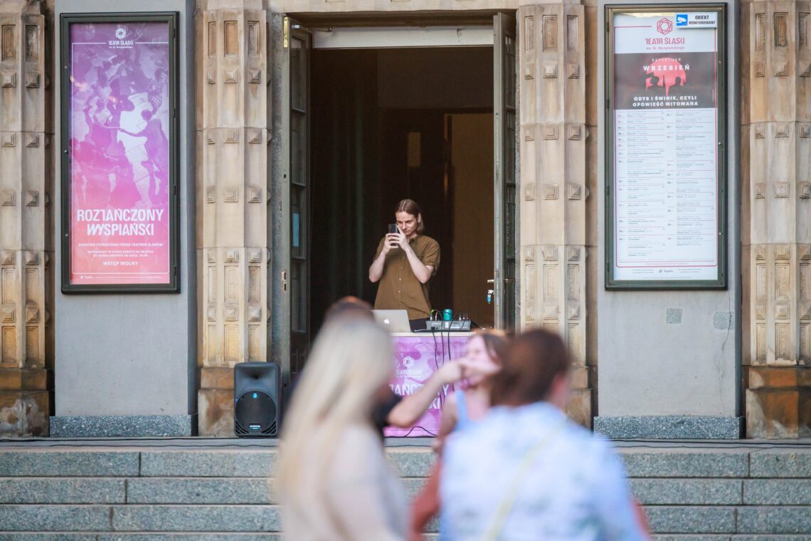 W otwartych drzwiach Teatru stojący przy sprzęcie muzycznym mężczyzna w trakcie pracy, na ścianie teatru w gablotach z lewej strony plakat "Roztańczony Wyspiański, z prawej repertuar na wrzesień 2023