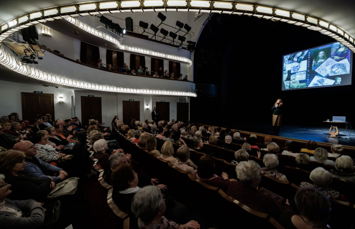 Ludzie siedzące na widowni Dużej Sceny Teatru Śląskiego, na scenie kobieta przemawiająca do mikrofonu, za nią wyświetlony na zawieszonym ekranie slajd