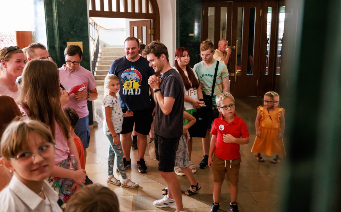 Grupa osób w różnym wieku w foyer Dużej Sceny Teatru Śląskiego, osoby w trakcie zwiedzania, grupę oprowadza aktor Dawid Ściupidro