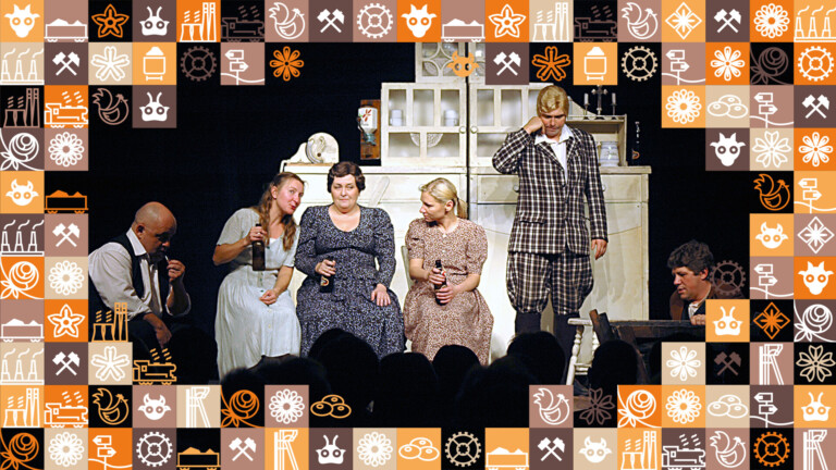 W centrum trzy kobiety siedzące na krzesłach, trzymają szklane butelki w dłoniach, koło nich mężczyźni - jeden siedzi obok, drugi stoi, trzeci siedzi przy stole; wokół zdjęcia ramka z małych kwadracików w różnych kolorach (odcienie pomarańczowego i brązowego) przedstawiające obrazki związane ze Śląskiem, np. szyb górniczy, wagon z węglem, kluski śląskie