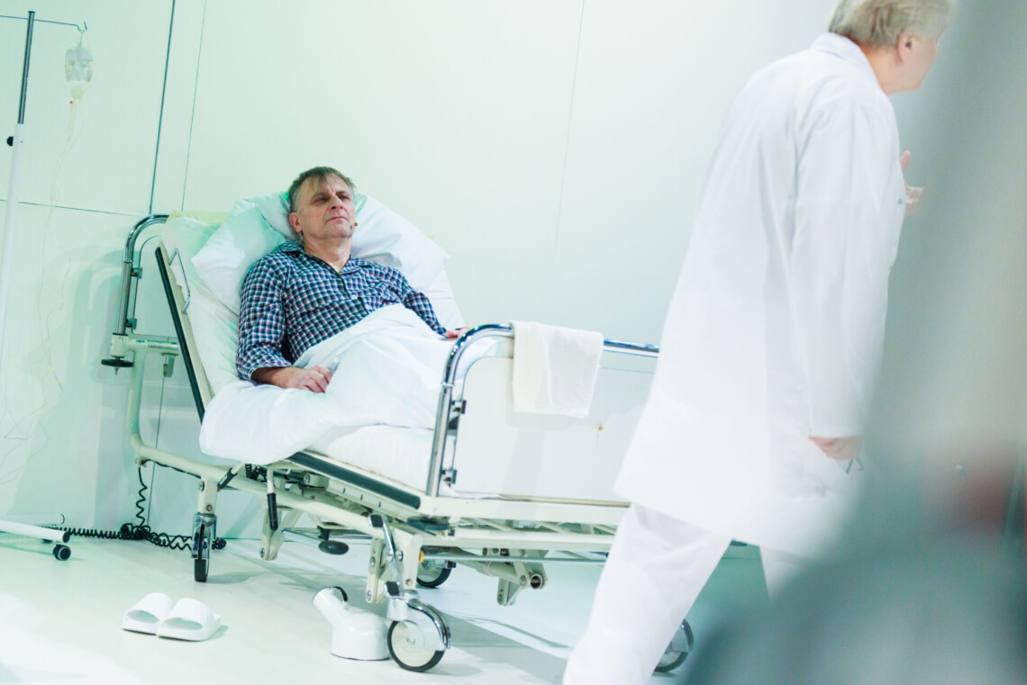 Mężczyzna siedzący w łóżku szpitalnym, za nim biała ściana, przed łóżkiem przechadza się mężczyzna ubrany na biało