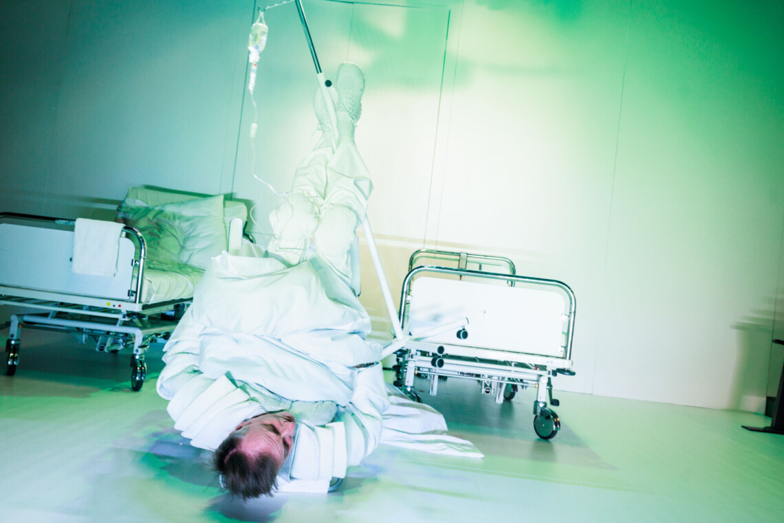 Mężczyzna ubrany na biało leżący na plecach na podłodze, między nogami przytrzymuje metalowy stojak z kroplówką, za nim dwa łóżka szpitalne, wszędzie panuje biel, scena oświetlona na zielono