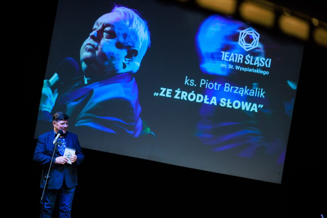 Dyrektor Robert Talarczyk stoi przy mikrofonie na statywie, w ręce trzyma książkę, za nim ekran z wyświetlonym plakatem ze zdjęciem ks. Piotra Brząkalika promującym książkę "Ze źródła słowa"