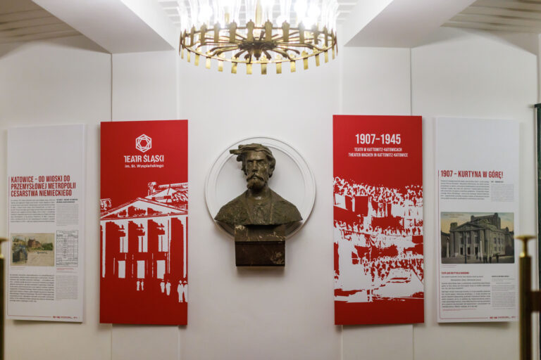 Cztery plansze wystawy "1907-1945. Teatr w Kattowitz-Katowicach", białe plansze po bokach z informacją, po środku czerwone plansze z białymi rycinami z lewej budynku Teatru, z prawej widowni, pomiędzy rycinami rzeźba z brązu - popiersie Stefana Wyspiańskiego