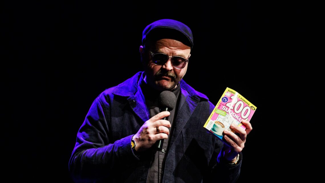 Stojący mężczyzna w przyciemnianych okularach, w lewej ręce trzyma książeczkę 200 sudoku, w prawej ręce trzyma mikrofon