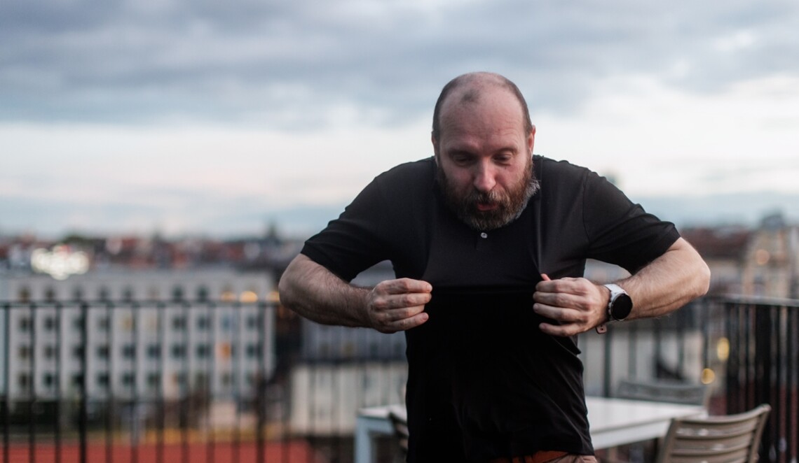 Mężczyzna stojący na balkonie, ubrany w czarna koszulkę polo, dłońmi naciąga koszulkę przed siebie odsuwając ją od ciała, patrzy w dół, za nim w tle widoczne miasto