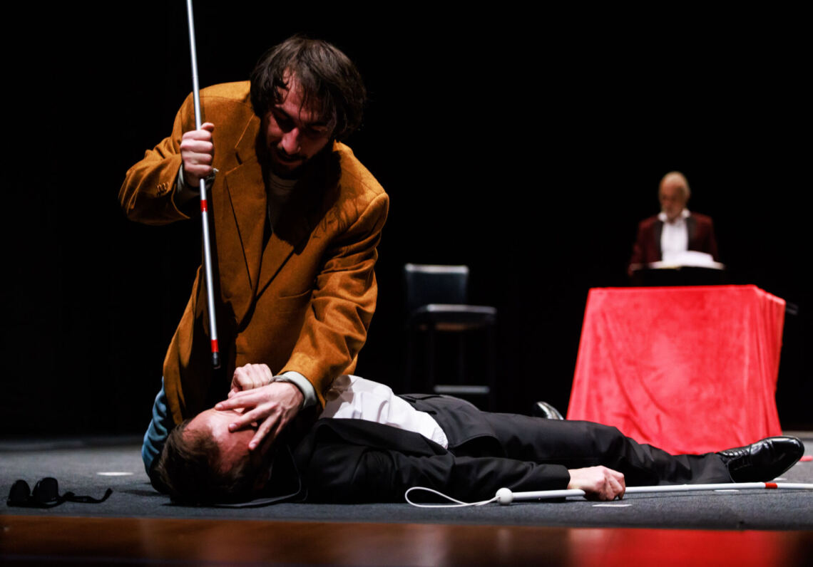 A man in a black suit lies on his back on the floor, holding a white cane in his hand, a man in an orange jacket kneels over the lying man in suit, he touches his the eyes, he holds his white cane above his head, it looks like he is aiming at his eyes, in the background a man in a burgundy jacket stands behind a table with a red tablecloth