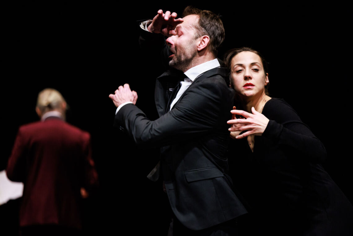 A man and a woman dressed in black standing sideways, the man has his eyes closed, his hand is on his forehead, he is looking forward, the woman is leaning against the man, she is looking sideways, towards the camera, she is holding the fingers of both hands together with their crests