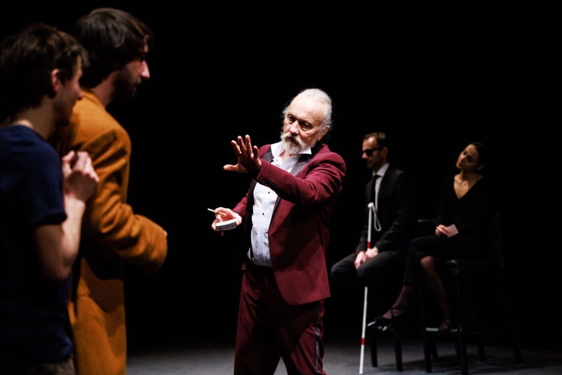 In the center stands a man in a burgundy suit, his left hand is stretched forward, in front of him stand two men, behind him sit a man and a woman dressed in black on high chairs, the man has black glasses, he holds a white cane in his hand