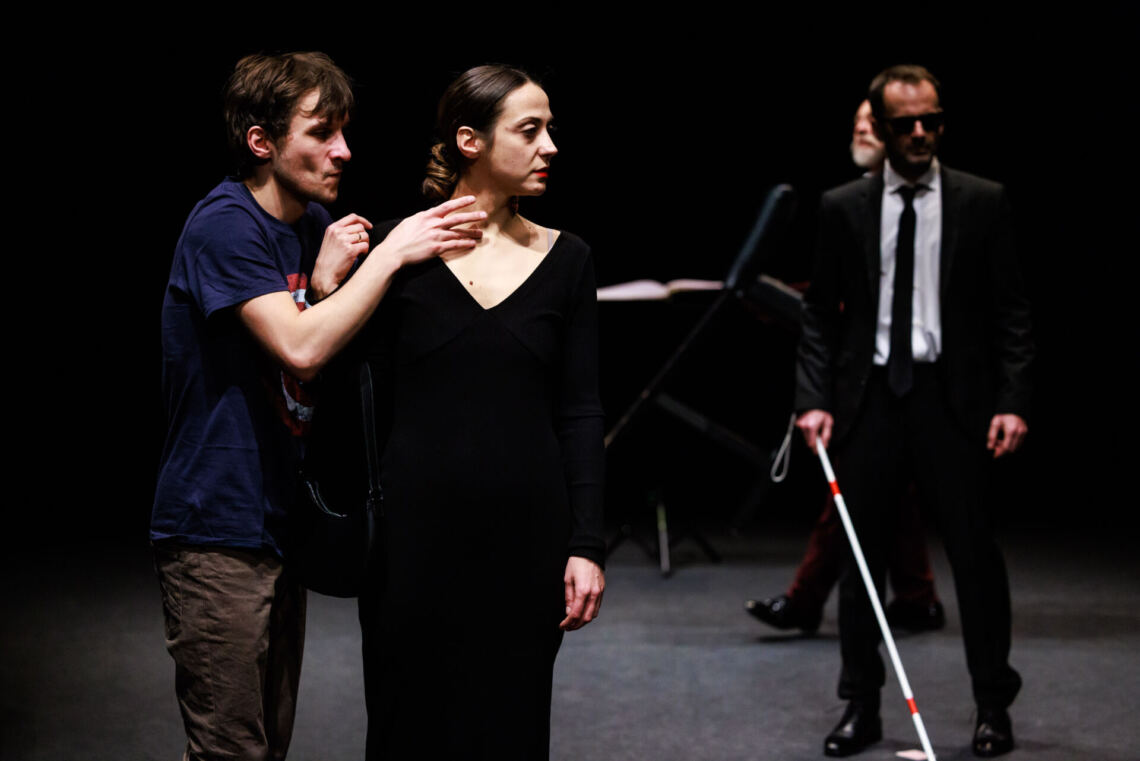 A man in a t-shirt touches the neck of a woman wearing a black dress with a low neckline, the man is standing sideways, the woman's head is turned in the opposite direction, in the background a man in a black suit with a white cane is visible