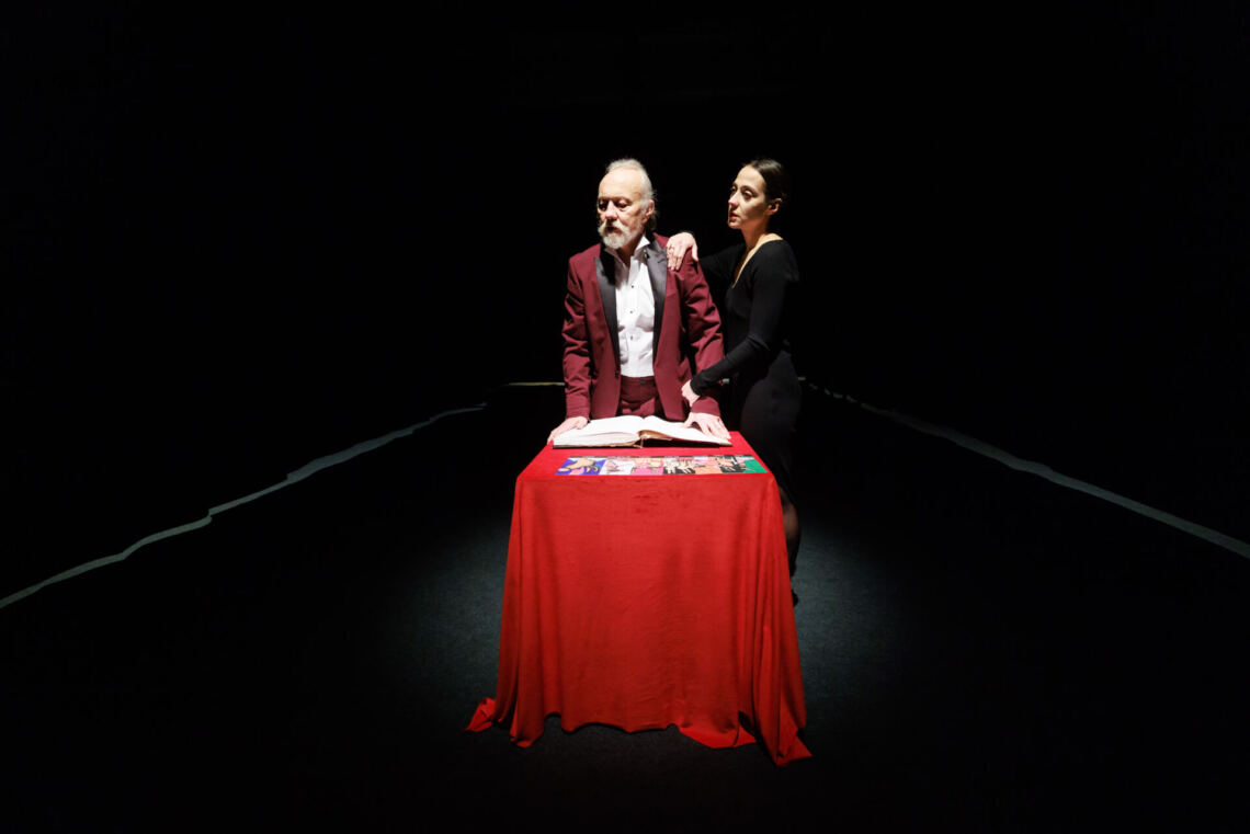 At a table with a red tablecloth, on which lie colorful cards and a book, stands a man in a burgundy jacket, next to which stands a woman dressed in black
