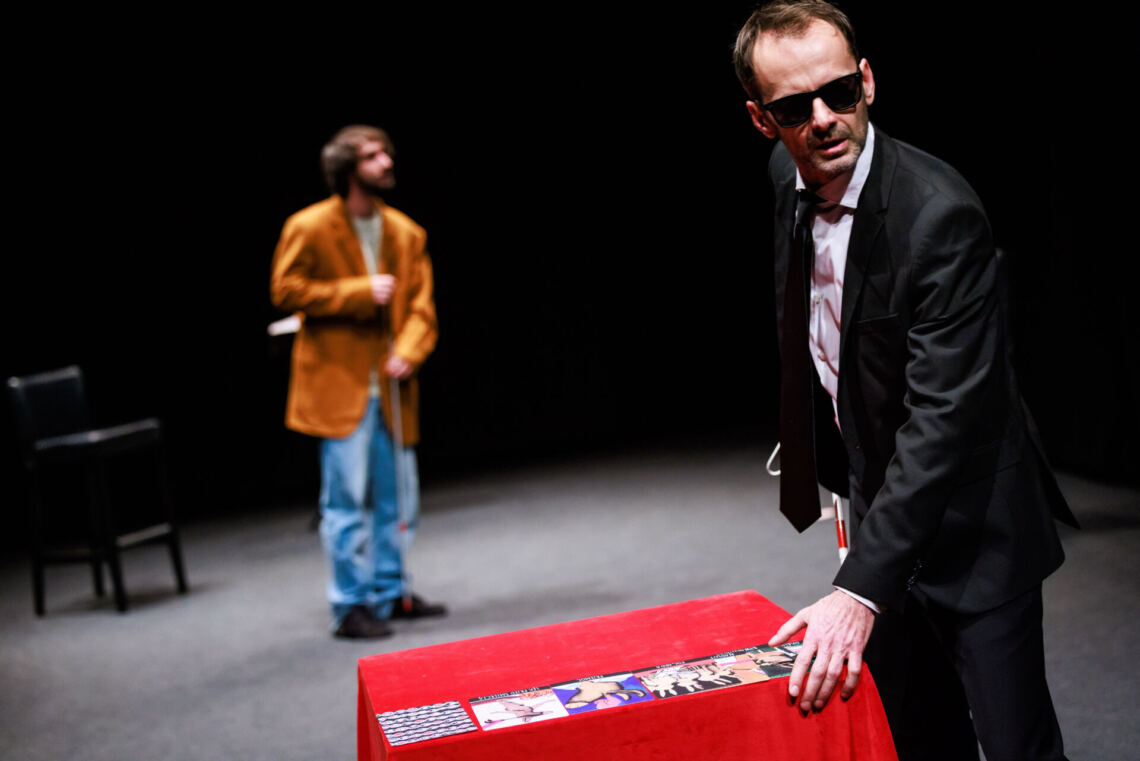 At the red table stands a man in a black suit and black glasses, on the table there are colorful cards, in the background there is a man in an orange jacket