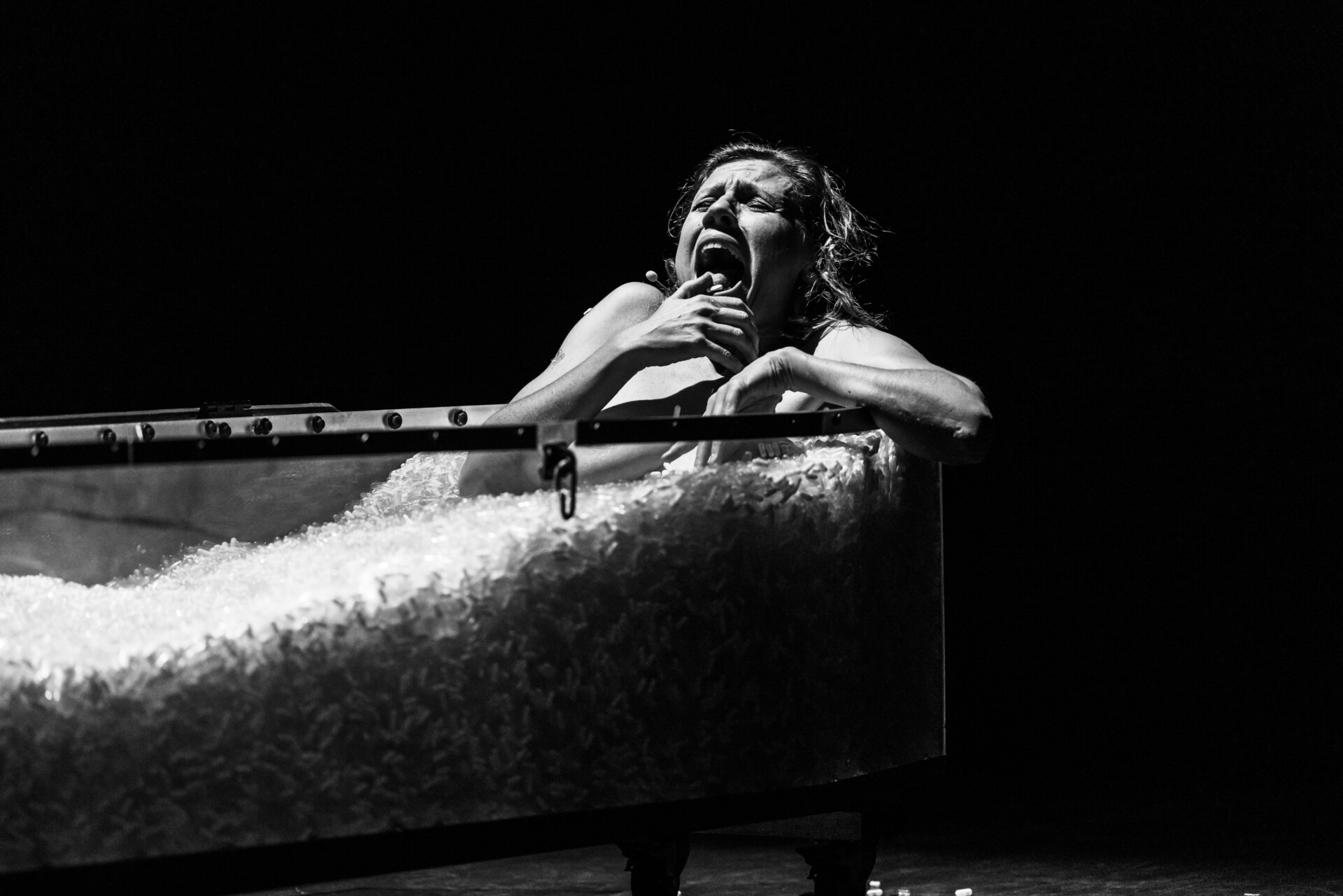 Naga kobieta leży w szklanej trumnie wypełnionej kapsułkami z lekami. Kciuk prawej dłoni wtrzyma w otwartej buzi. Zdęcie czarno-białe.