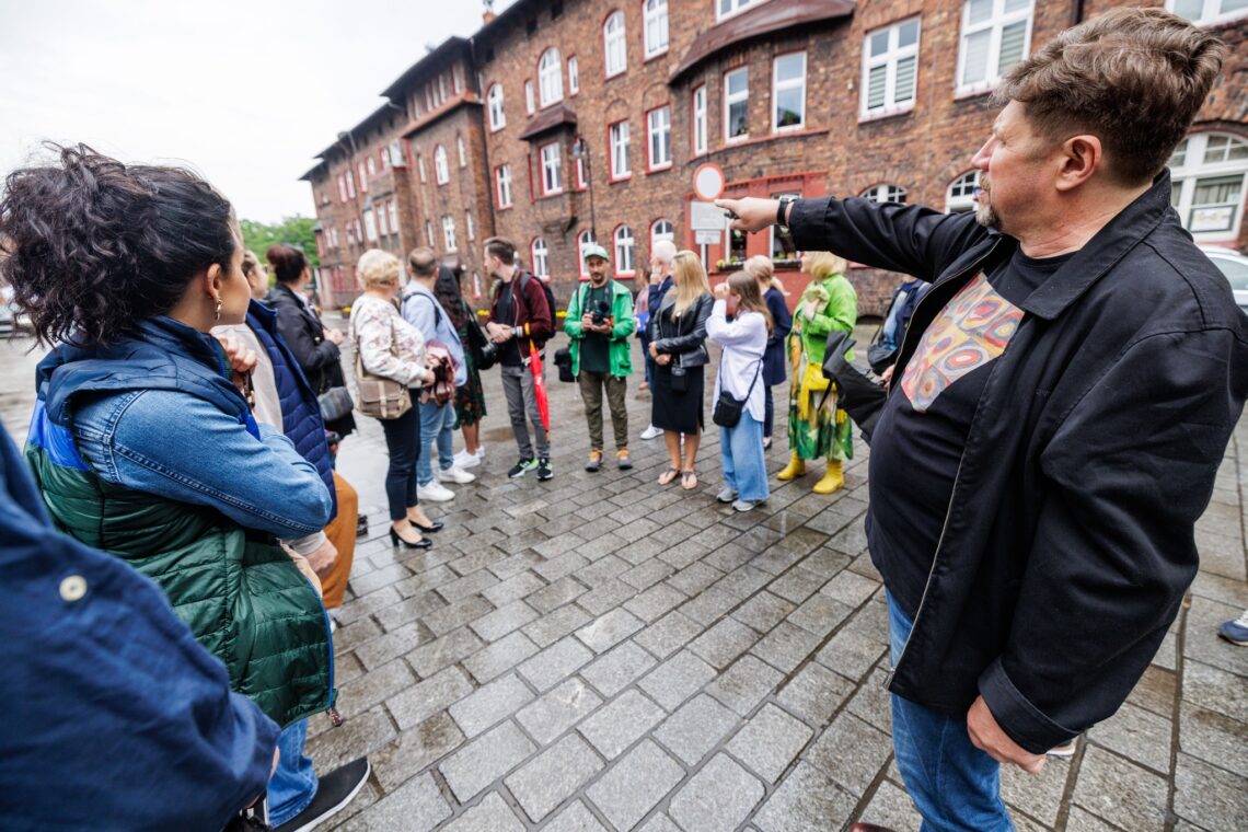 Dyroktor Robert Talarczyk z wyciągniętą w przód prawą ręką oraz wskazuzjący palec, pokazuje coś w oddali, osoby stojące przed nim w półkolu patrzą w tamtym kierunku, odwracają się do tyłu