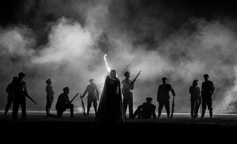 W rzędzie stoją osoby z długą bronią w ręce, w centrum wyróżnia się jedna postać, widoczne są jedynie zarysy postaci, za nimi dym; zdjęcie czarno-białe