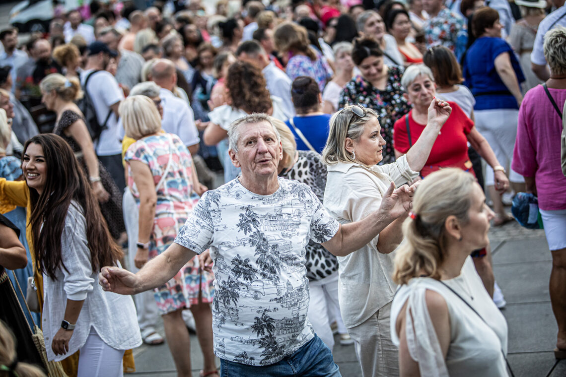 Mężczyzna w trakcie tańca z rozłożonymi na bok rękami, wokół niej tańczące osoby