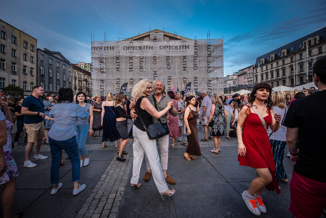 W centrum kobieta i mężczyzna tańczący w parze, wokół nich inne osoby, w tle widoczny zasłonięty budynek Teatru w trakcie remontu