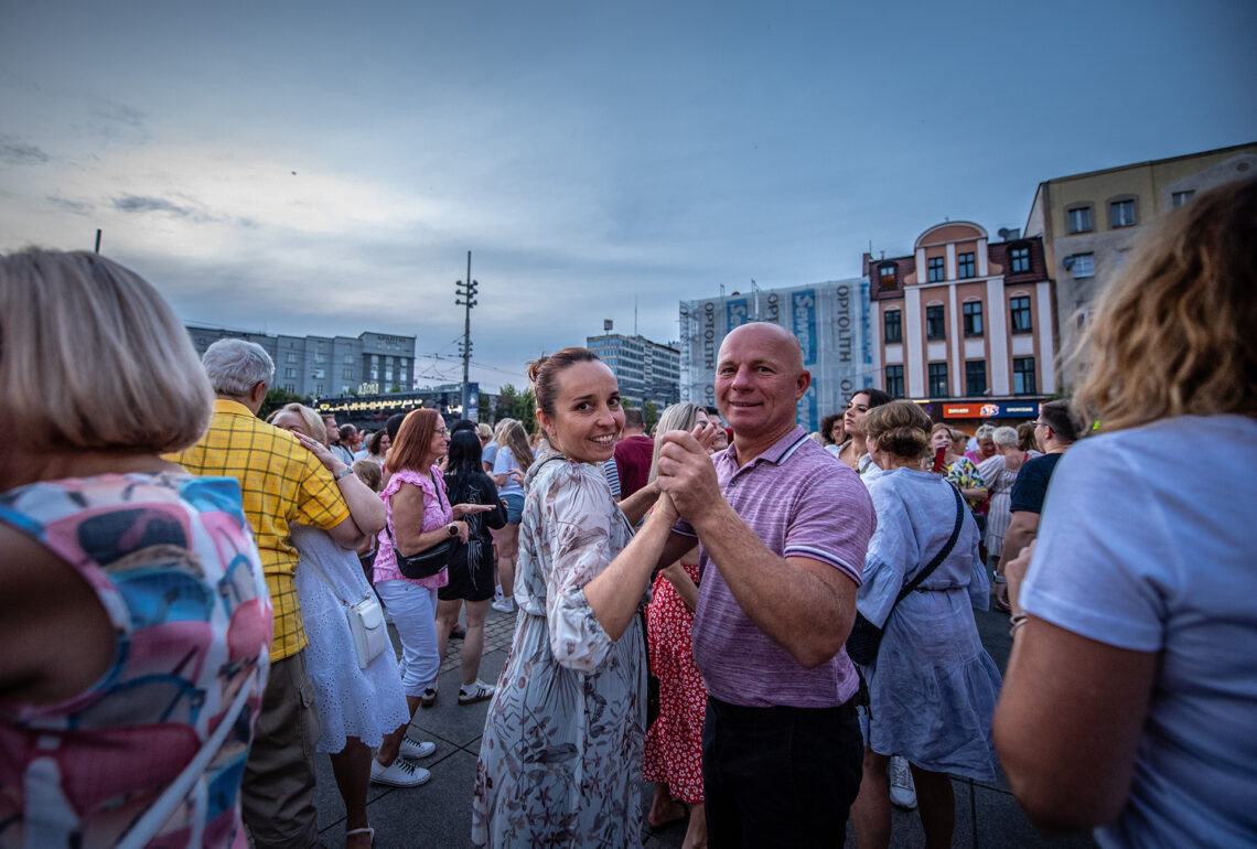 Kobieta i mężczyzna tańczący w parze, patrzą się w obiektyw, wokół nich inne osoby