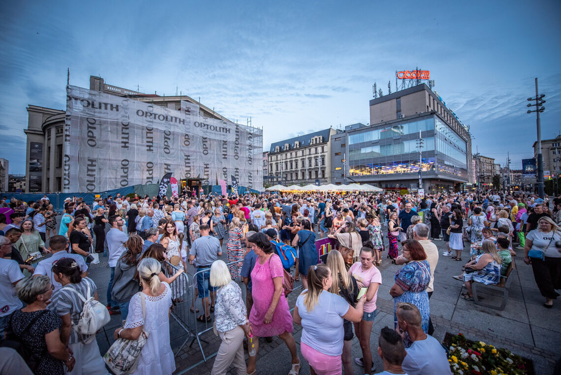 Plac przed Teatrem wypełniony ludźmi, w tle widoczne miasto, w tym zasłonięty budynek teatru w trakcie remontu, ujęcie z góry