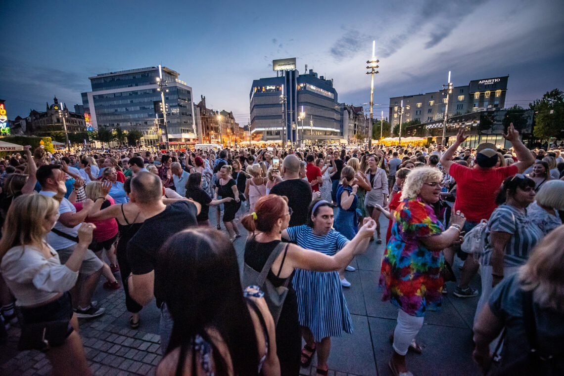 Tłum osób w trakcie tańca na placu, w ple widoczne miasto nocą