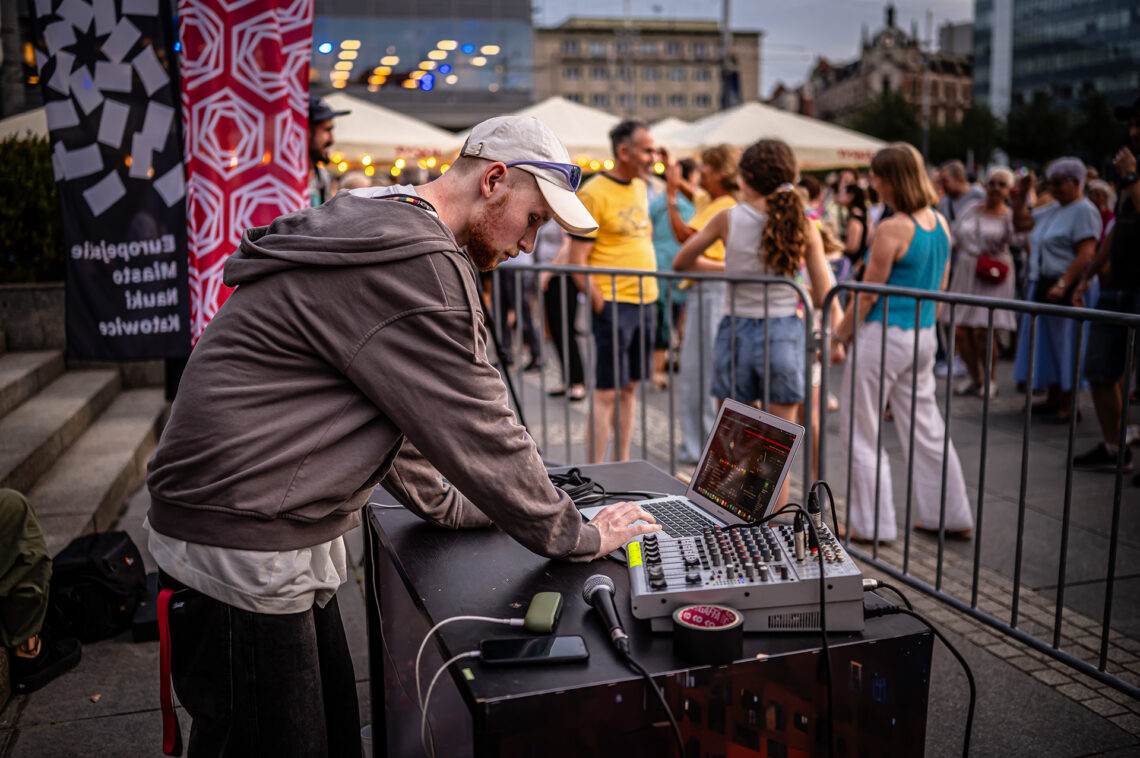 Mężczyzna obsługujący muzykę przy pracy na komputerze stojącym na stole, obok stoi mikser dźwiękowy