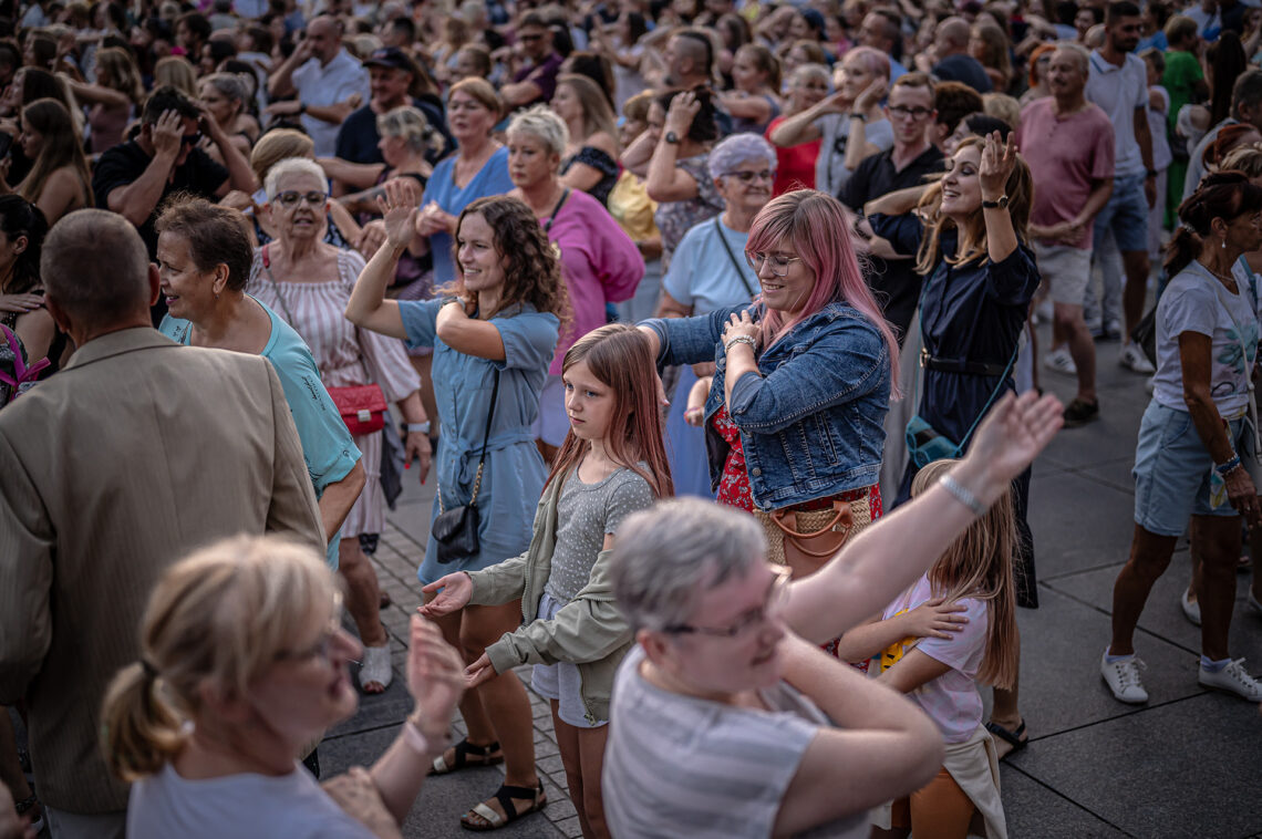 Tłum osób na placu, większość trzyma jedną dłoń na ramieniu lub na głowie - kroki popularnego tańca macarena