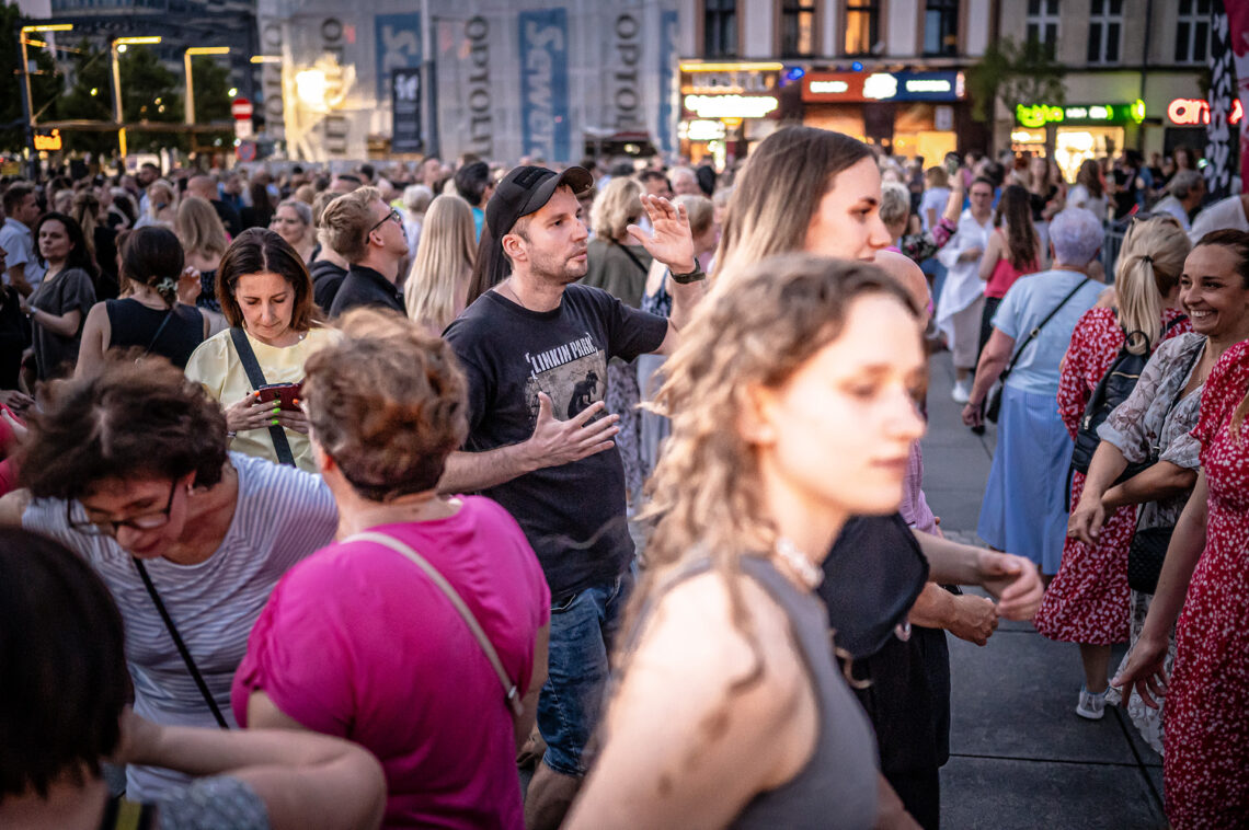 Tłum osób na placu, w centrum mężczyzna w czarnej czapce uchwycony w trakcie tańca, prawą rękę ma wysuniętą do przodu, lewą ku górze