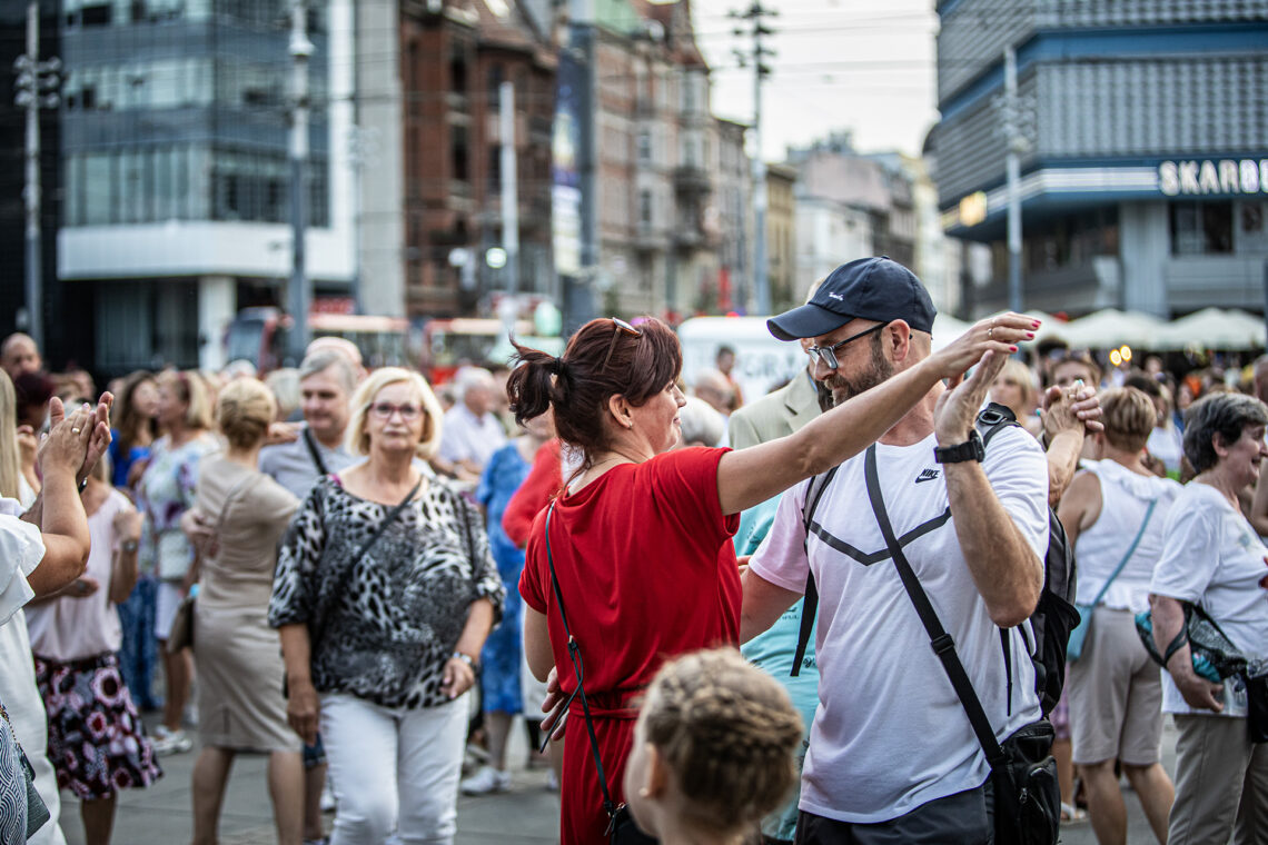 Kobieta ubrana na czerwono i mężczyzna tańczący w parze, za nimi inne osoby