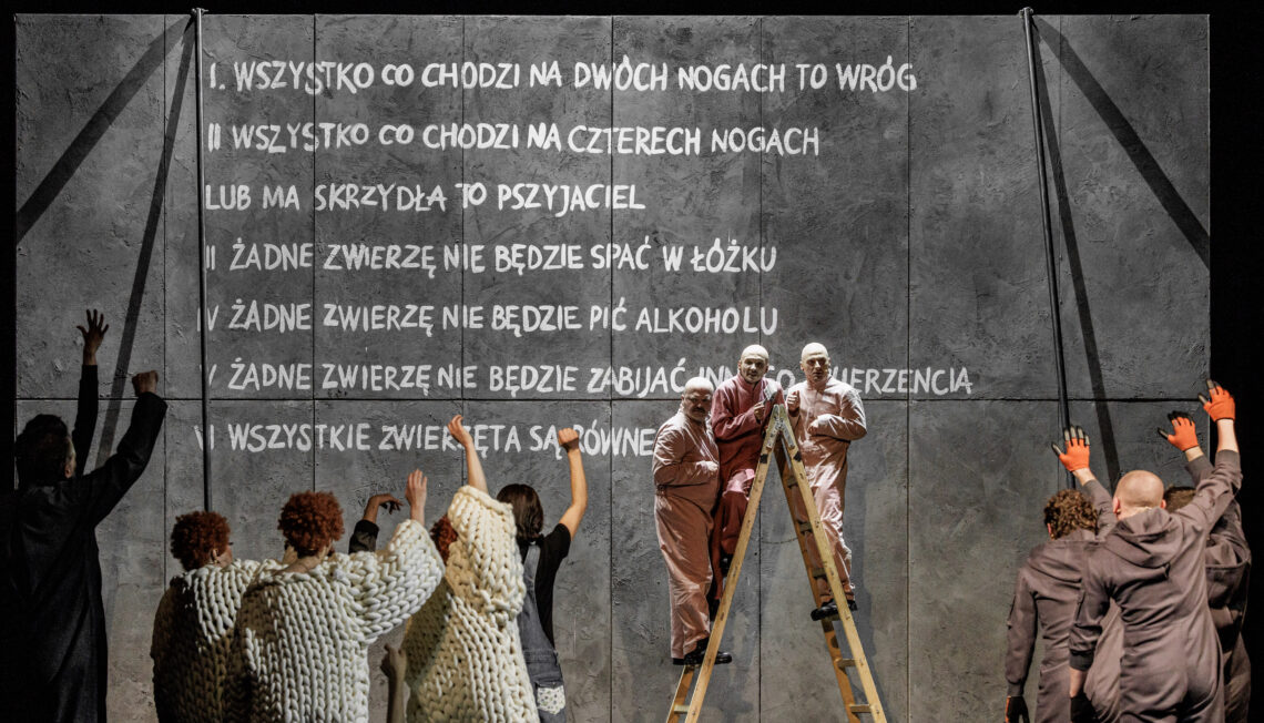 Na pierwszym planie tyłem w grupach stoją postacie ubrane w takie same stroje, pod ścianie na drabinie przodem stoją trzy postacie w różowych strojach, za nimi szara ściana z wypisanymi przykazaniami