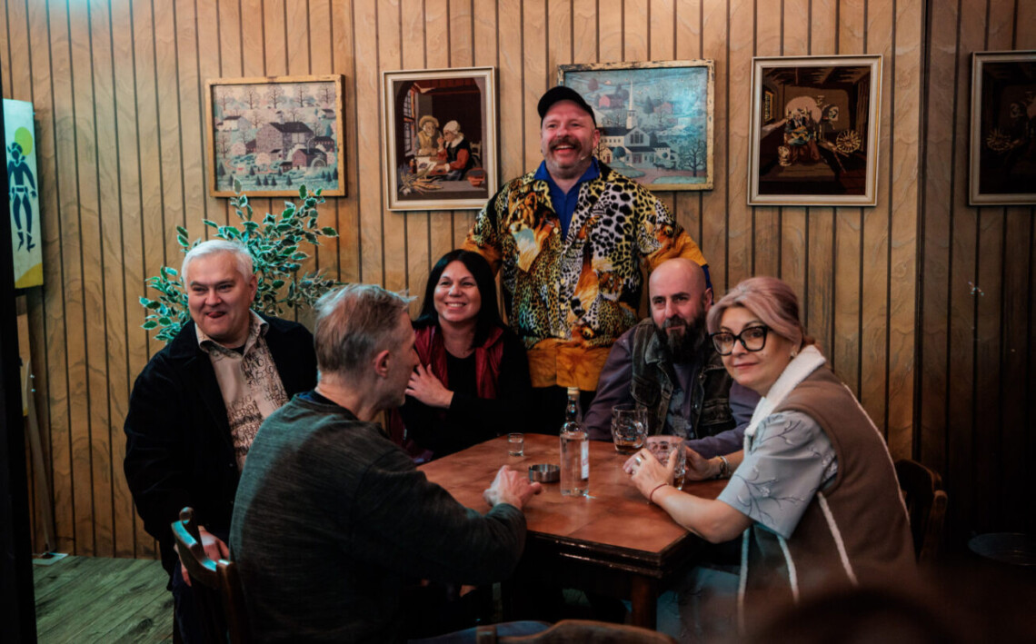 Pięć osób siedzących za stołem, między nimi przy ścianie stoi mężczyzna, na stole stoi butelka wódki