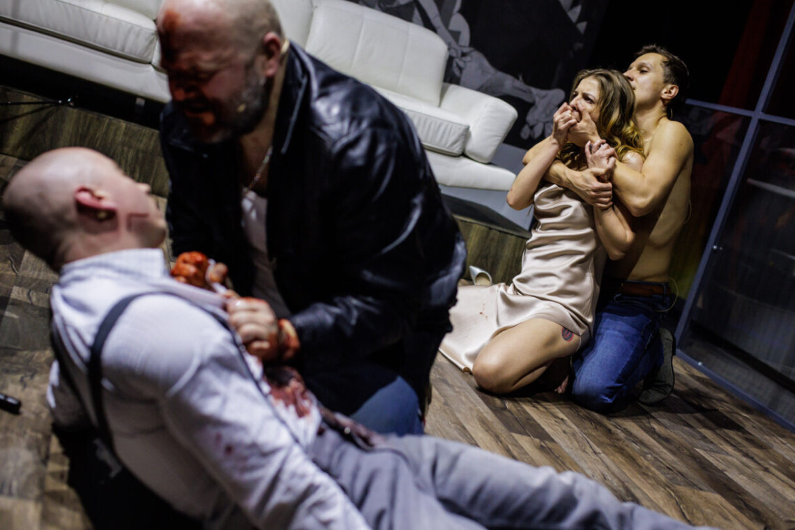 Na pierwszym planie rozmazana postać dwóch mężczyzn - mężczyzna klęczy nad mężczyzną z zakrwawioną koszulą, na drugim planie klęczące osoby - mężczyzna bez koszulki trzyma od tyłu kobietę i zasłania jej usta dłonią