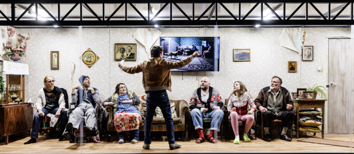 Osoby siedzące w rzędzie pod ścianą w pokoju, fotel po środku jest pusty, przed nim stoi mężczyzna z uniesionymi ku górze rękami, mężczyzna jest zwrócony twarzą do siedzących osób