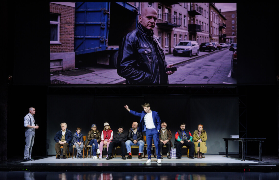 Na krzesłach w rzędzie siedzą różne osoby, przed nimi w centrum stoi mężczyzna w garniturze, z wyciągnięta w bok dłonią w kierunku mężczyzny stojącego z brzegu, nad nimi zawieszony ekran z projekcją, na którym widać mężczyznę na ulicy z domami z cegły